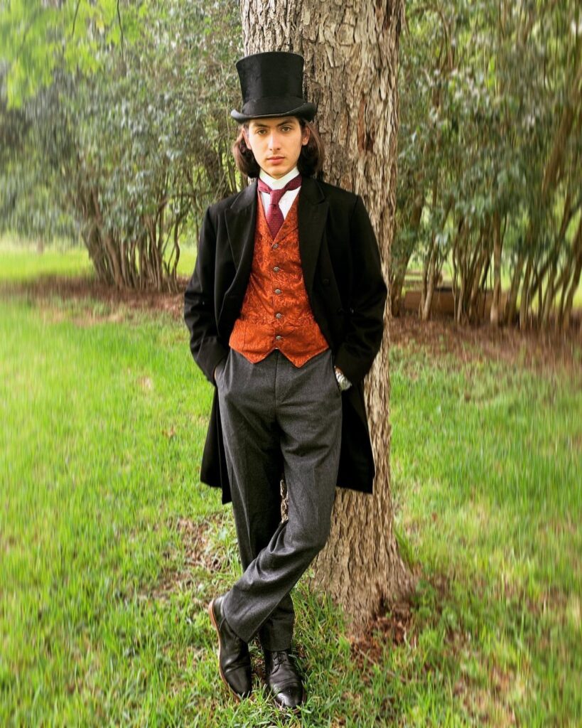 A young man in 1880s period clothing leans against a tree in a grassy field.