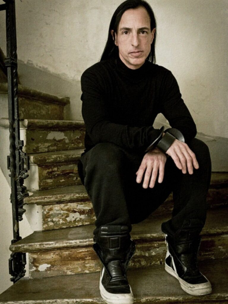 Man with long dark hair wearing black turtleneck and chunky sneakers sitting on wooden stairs.