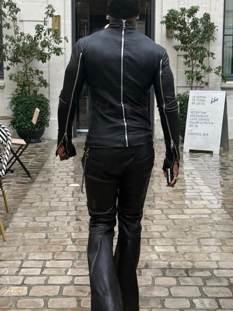 A person in tight black leather clothing walking through a cobblestone courtyard towards a building.