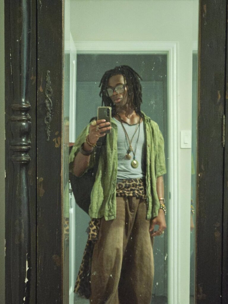 Man with dreadlocks wearing boho style clothing posing for a mirror selfie.