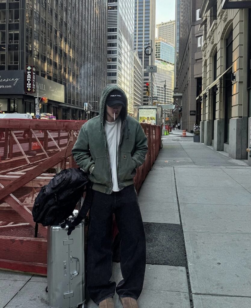 Person in a green hooded jacket, white shirt, and black pants standing on a city sidewalk.