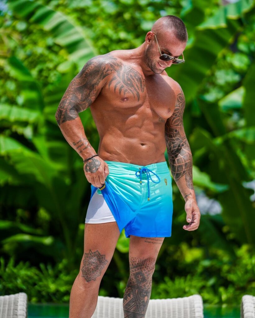 A muscular tattooed man wearing blue and white gradient swim trunks in a tropical setting.