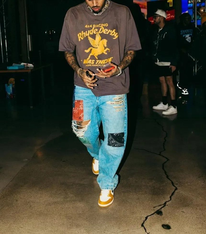 Man walking in a brown graphic t-shirt, patched distressed jeans, and yellow sneakers at night.
