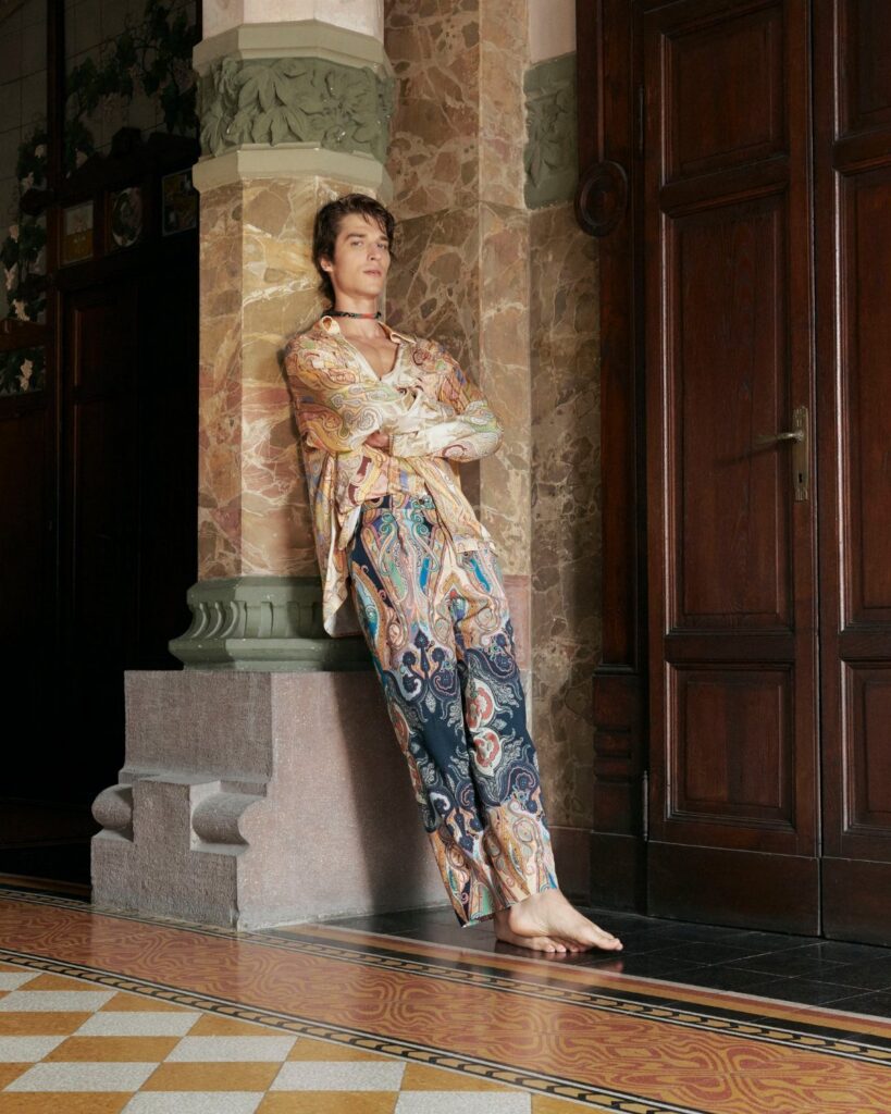A person wearing paisley patterned silk clothing leans against a marble pillar in an ornate room.