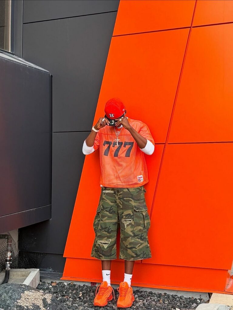 Man in orange shirt, camouflage shorts, and orange sneakers leaning against an orange wall.