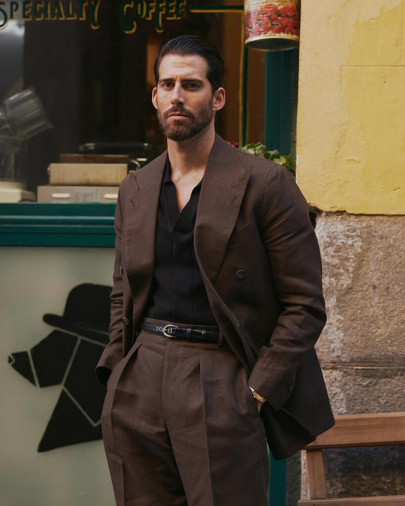 Man wearing a dark brown double-breasted suit and black shirt standing outdoors.