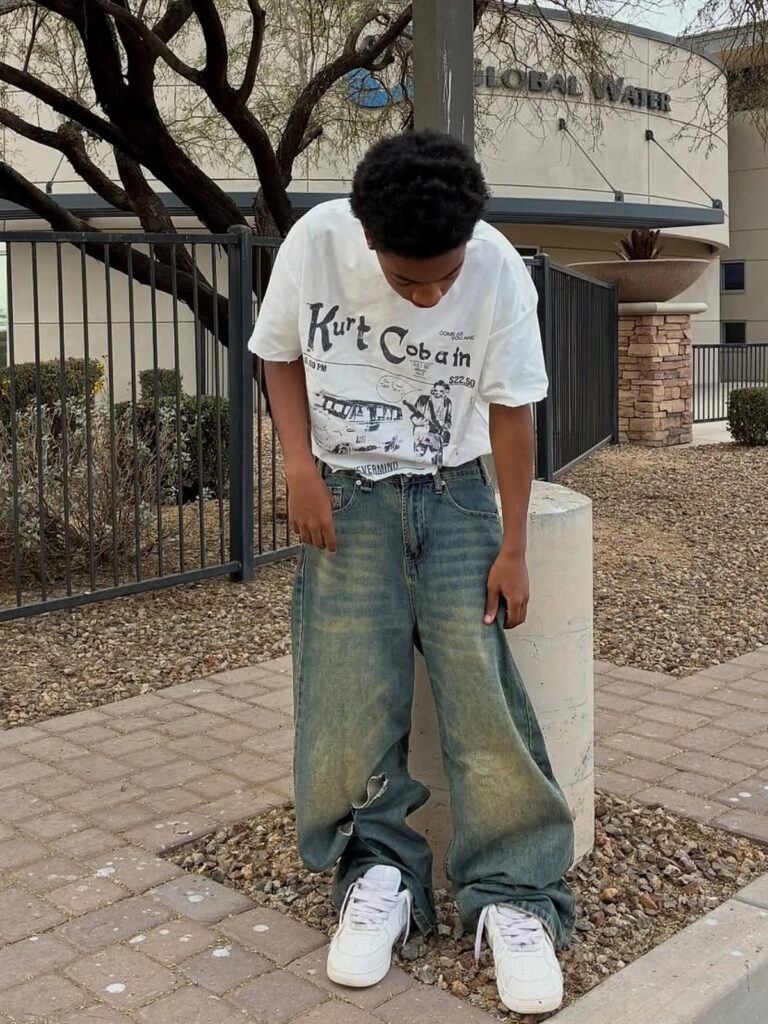A young man wearing a Kurt Cobain t-shirt, baggy jeans, and white sneakers standing outdoors.