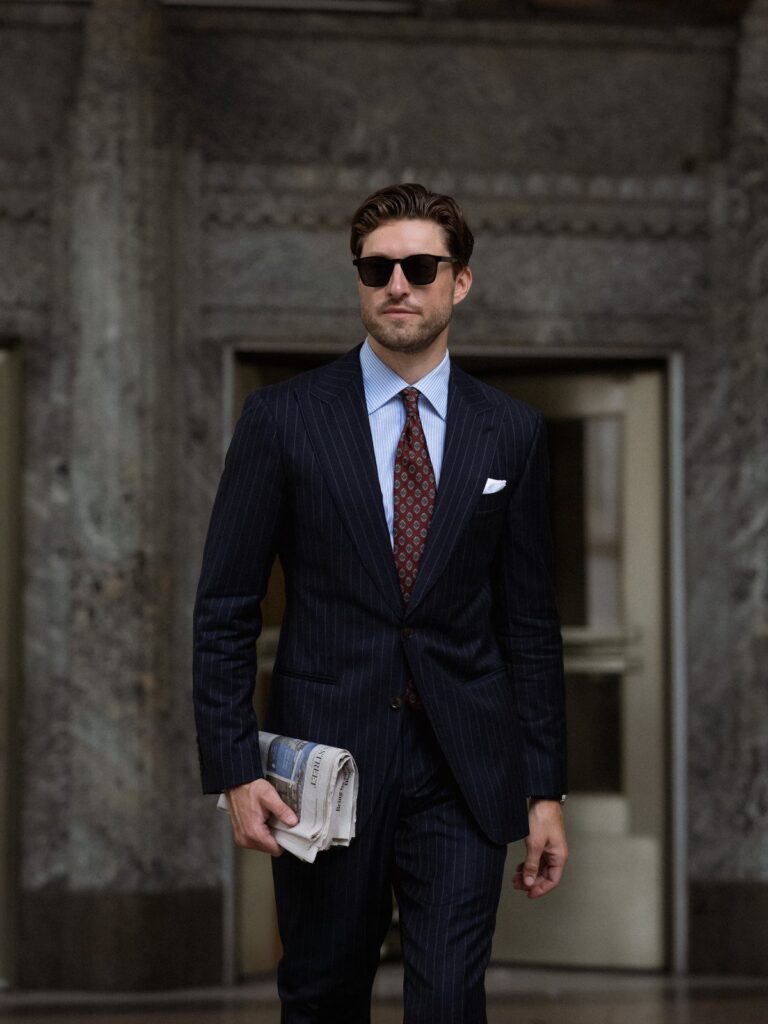 A man in a navy pinstriped suit, light blue shirt, and burgundy tie walking outdoors.
