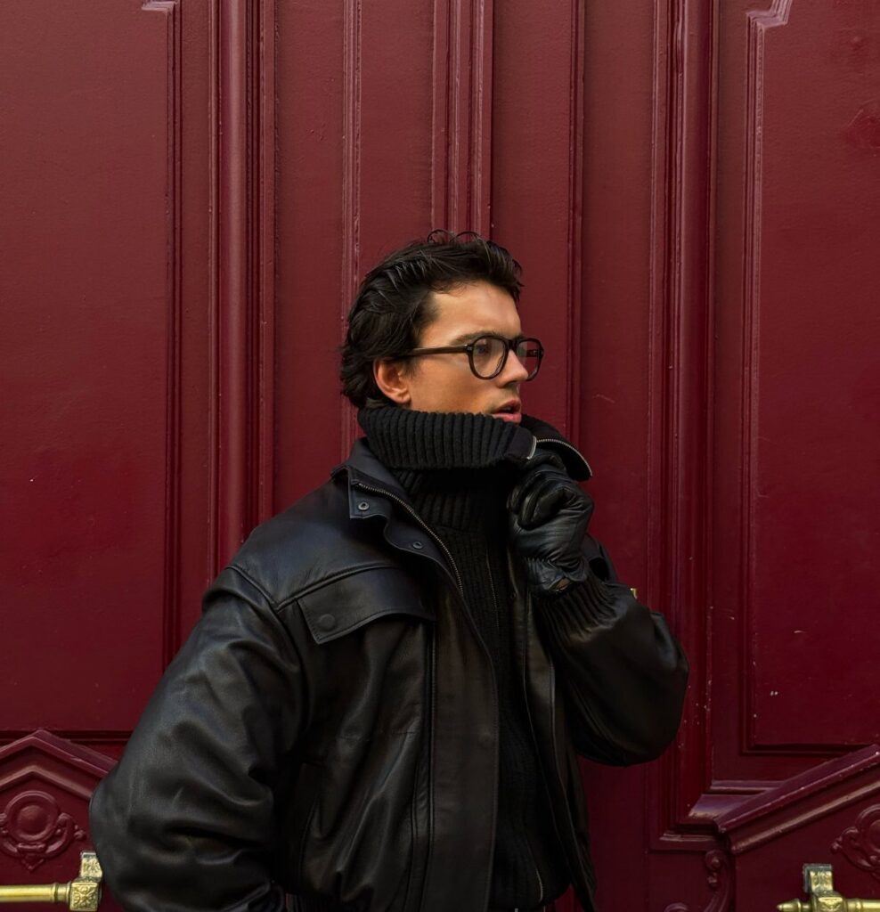A man wearing a black turtleneck and leather jacket against a red door.