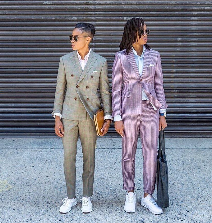 Two people posing in stylish, tailored suits against a grey corrugated metal background.