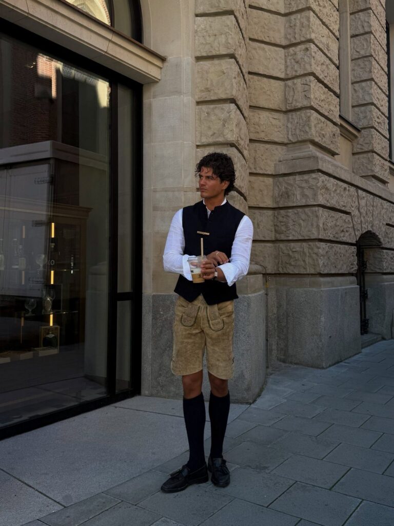 A man wearing traditional German leather shorts, a dark vest, and white shirt standing outdoors.
