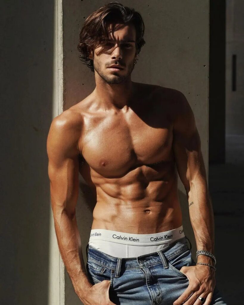 A muscular man with wavy hair posing shirtless against a concrete wall in dramatic lighting.