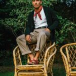 A man in a dark cardigan and khaki trousers sits on a bamboo chair outdoors.