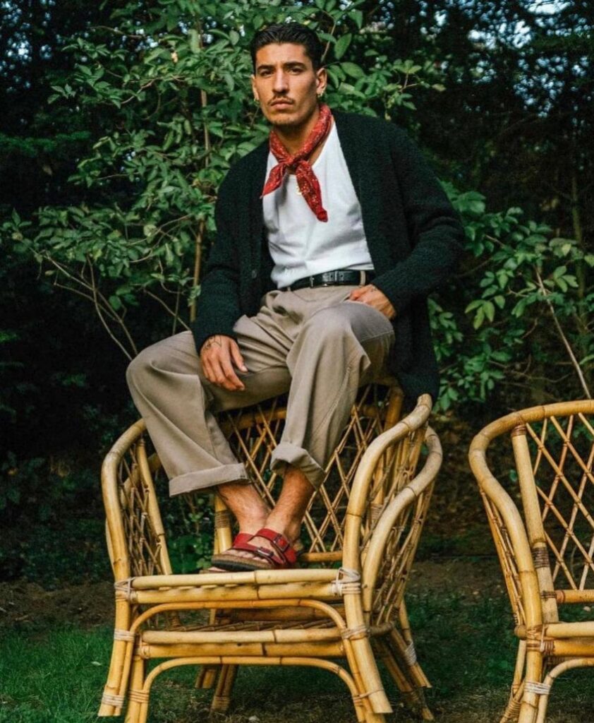 A man in a dark cardigan and khaki trousers sits on a bamboo chair outdoors.