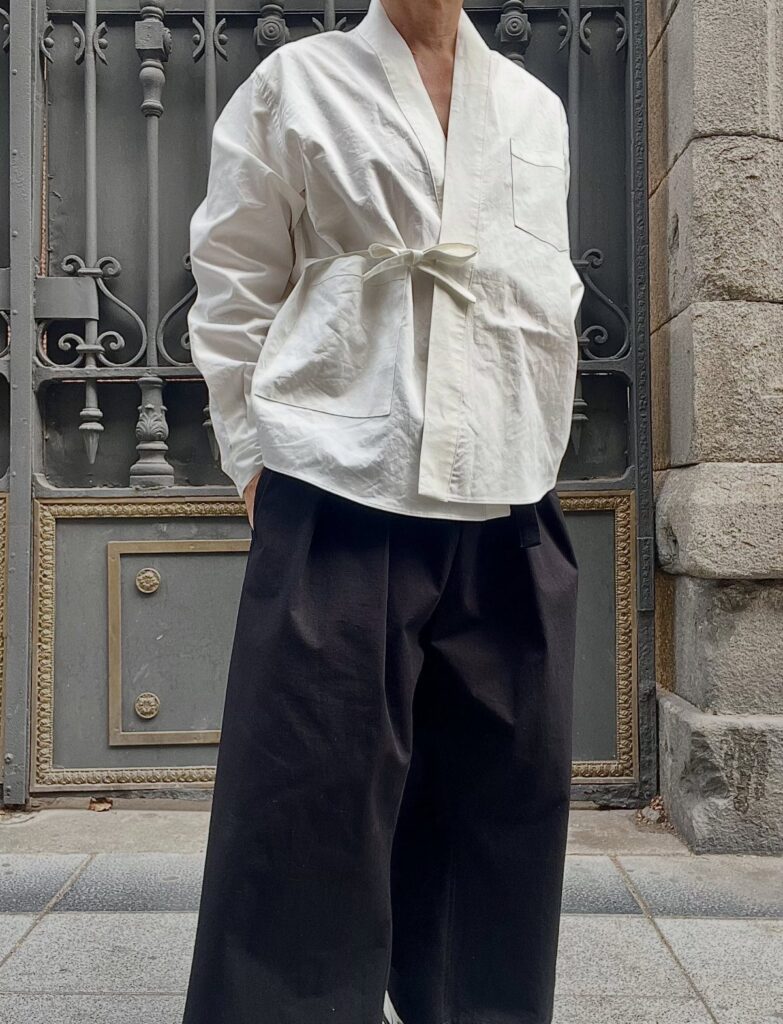 Person wearing a white wrap shirt and wide-leg black trousers standing by a metal gate.