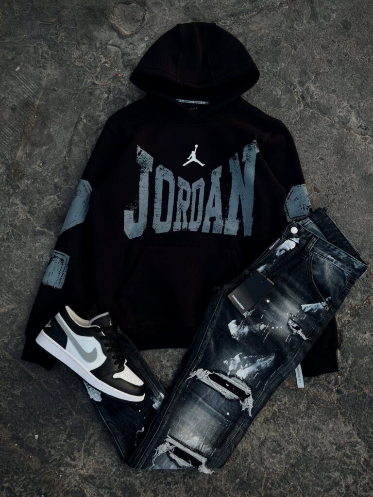 Black Jordan hoodie, distressed denim jeans, and black and white sneakers laid on concrete.