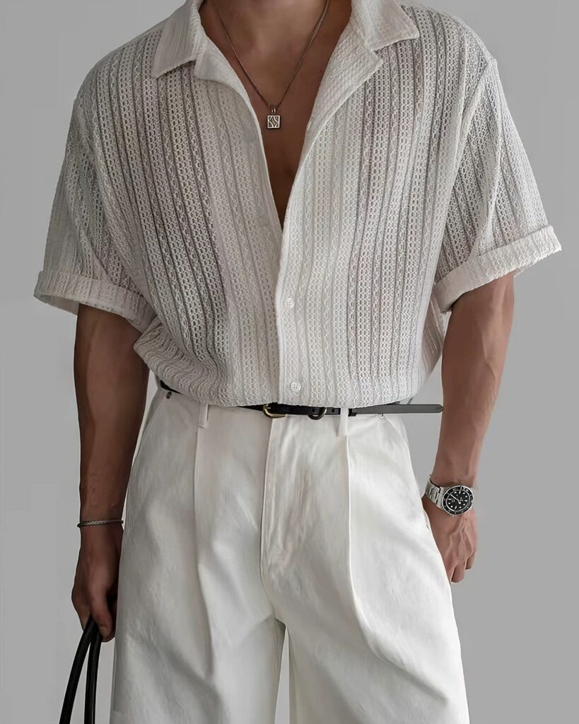 Man wearing a white textured mesh shirt and white pleated trousers with silver accessories.