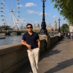 A man wearing a navy polo and cream trousers poses by the River Thames in London.