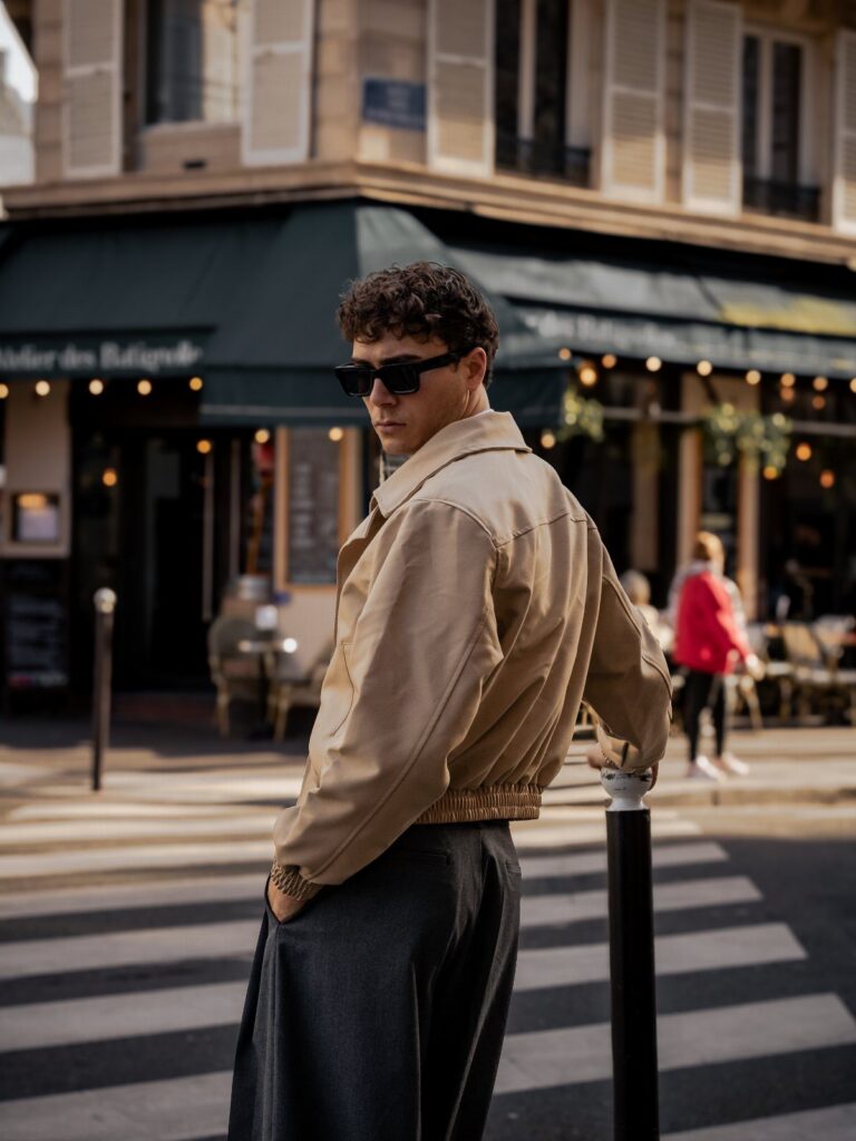 A young man in a tan jacket and sunglasses posing on a city street near a cafe.