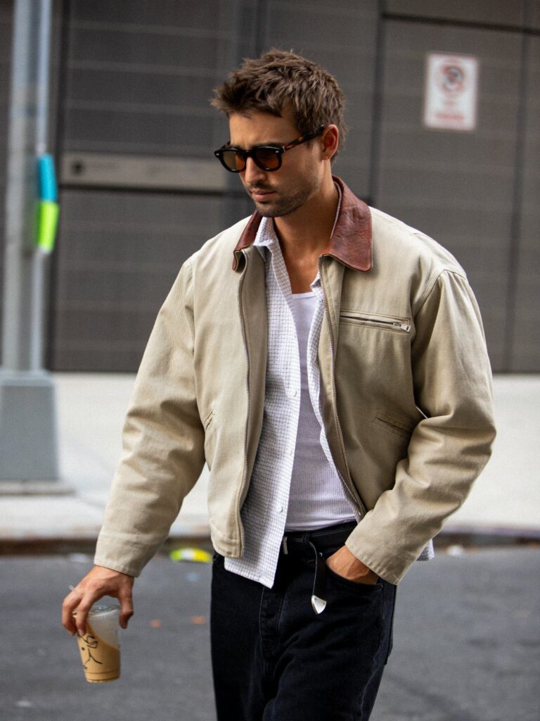 A man in a tan utility jacket and sunglasses walking down a city street.