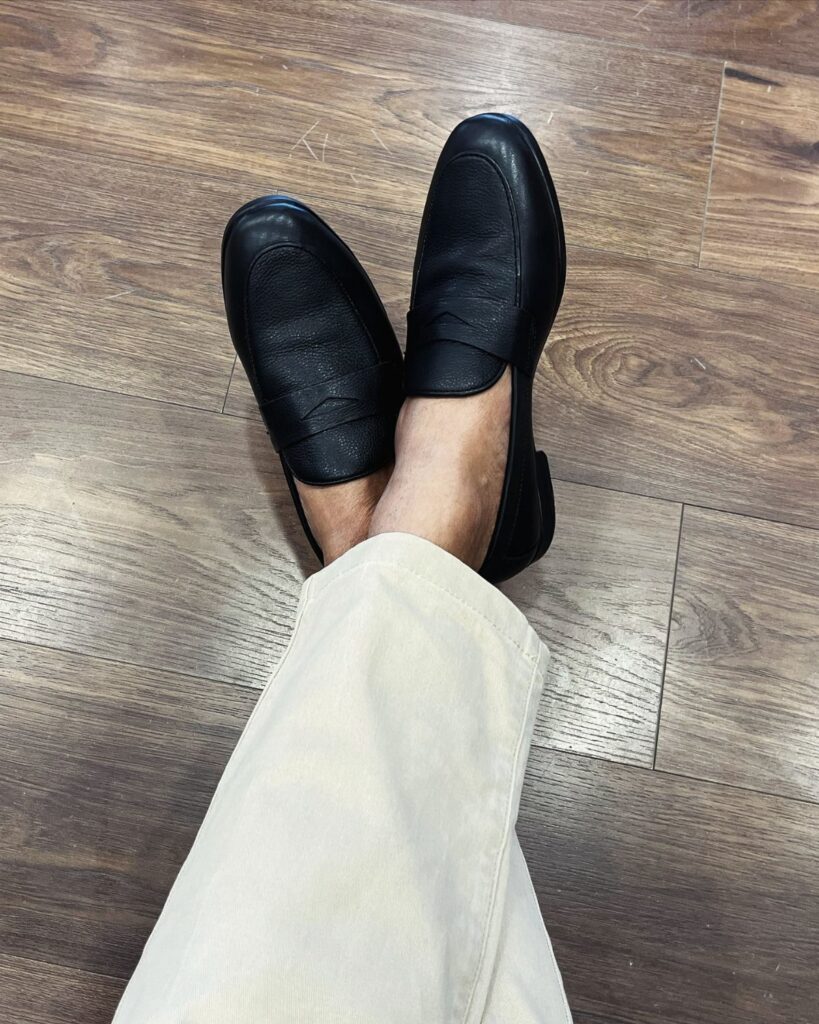 Black leather formal pump shoes worn with cream trousers on a wooden floor.