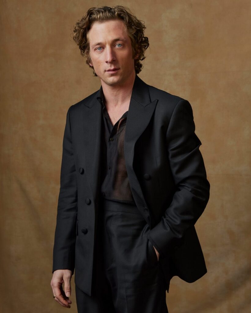 A man wearing a black blazer and a black sheer button-down shirt against a tan background.