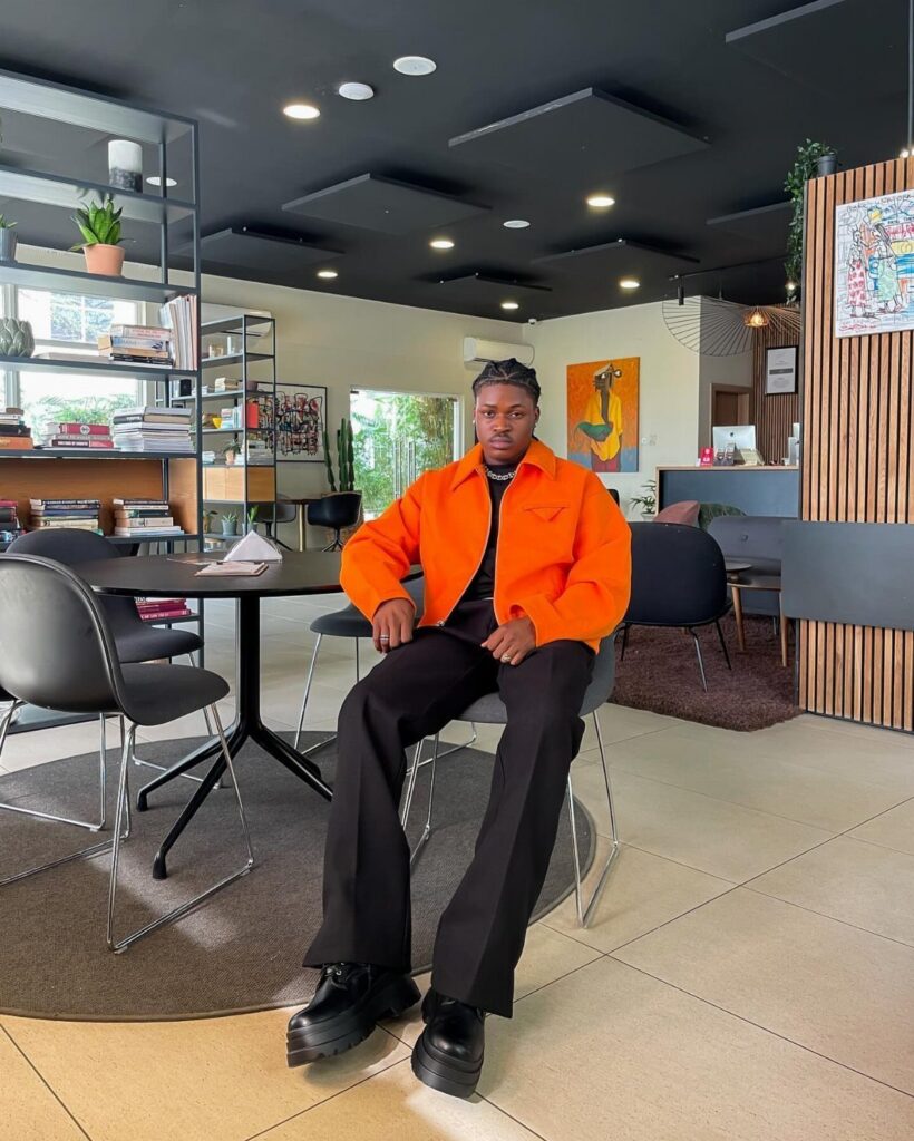 A man wearing an orange jacket, black pants, and chunky black combat boots sitting in a cafe.
