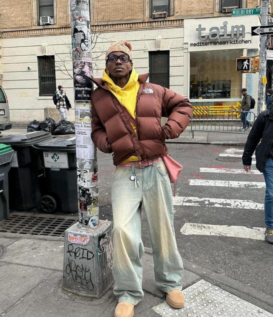 A man in a brown puffer jacket, yellow hoodie, and tan Ugg boots posing on a city street.