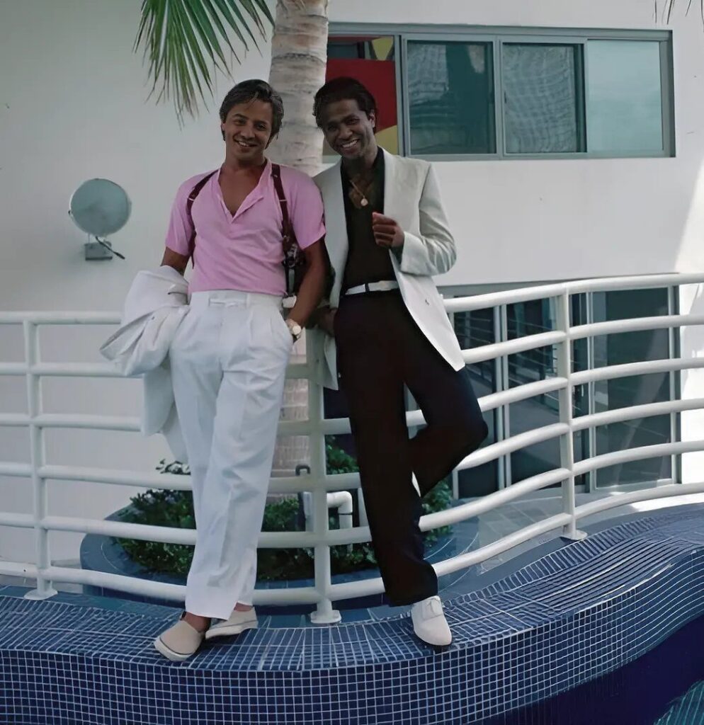 Two men posing in pastel and bright 1980s style clothing near a palm tree.
