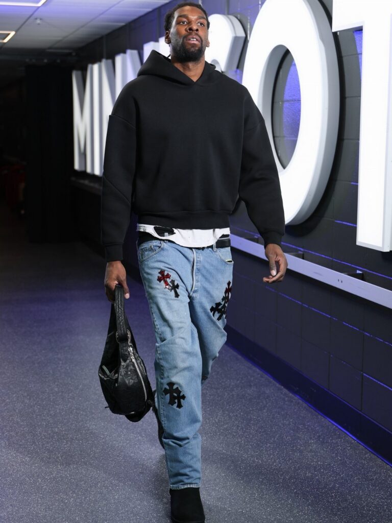 A man in a black hoodie and patterned jeans walks through a hallway carrying a bag.