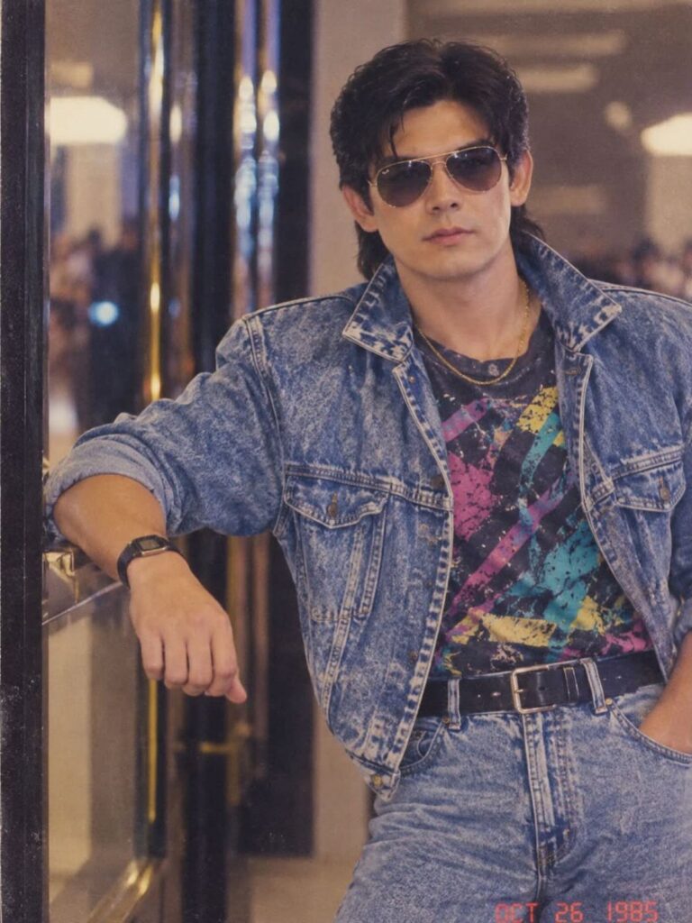 Man in sunglasses wearing an oversized denim jacket and colorful graphic t-shirt leaning against a door.