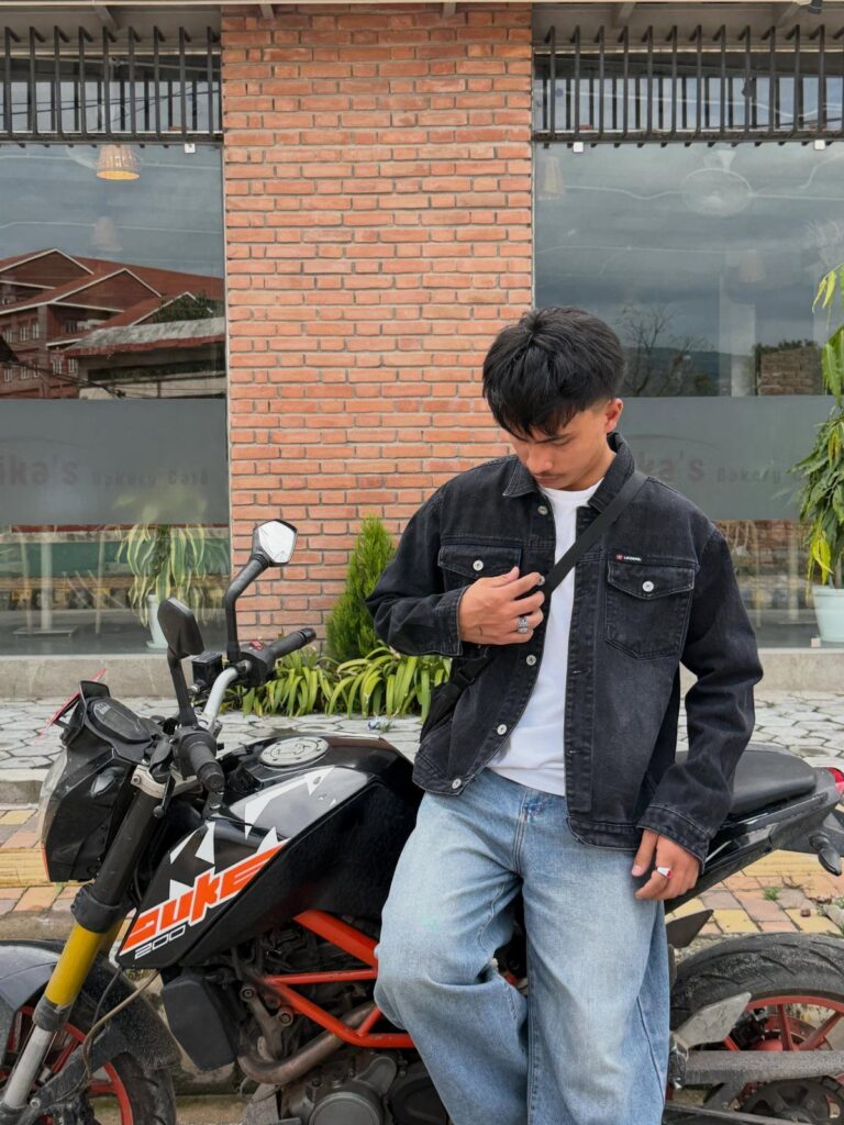 Young man in a black denim jacket and light blue jeans leaning on a motorcycle.