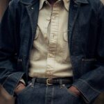 A person wearing a dark denim jacket, cream shirt, and blue denim jeans with a black belt.
