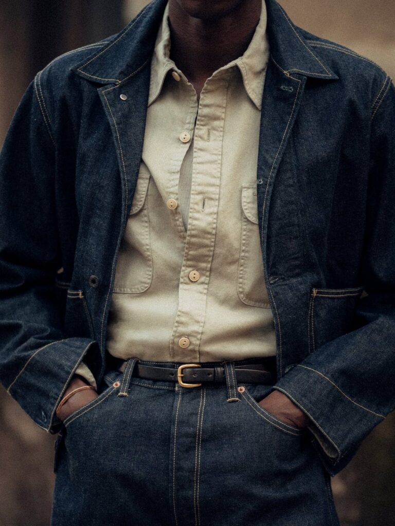 A person wearing a dark denim jacket, cream shirt, and blue denim jeans with a black belt.