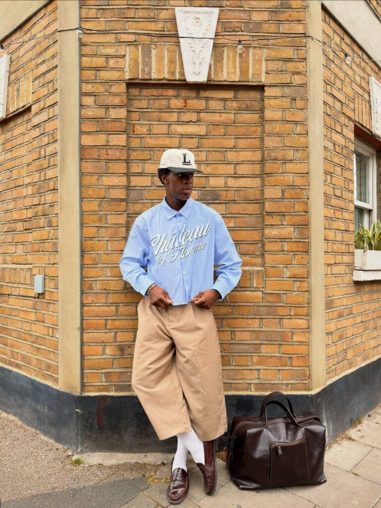 A man wearing a blue shirt and tan cropped wide-leg trousers standing by a brick wall.