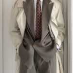 A person wearing a beige trench coat over a grey suit and patterned tie.