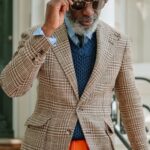 Man wearing a brown houndstooth blazer, navy sweater vest, and orange trousers while adjusting sunglasses.