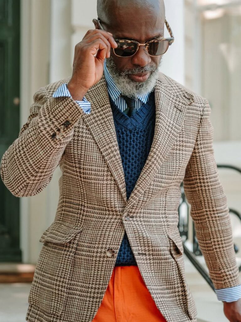 Man wearing a brown houndstooth blazer, navy sweater vest, and orange trousers while adjusting sunglasses.