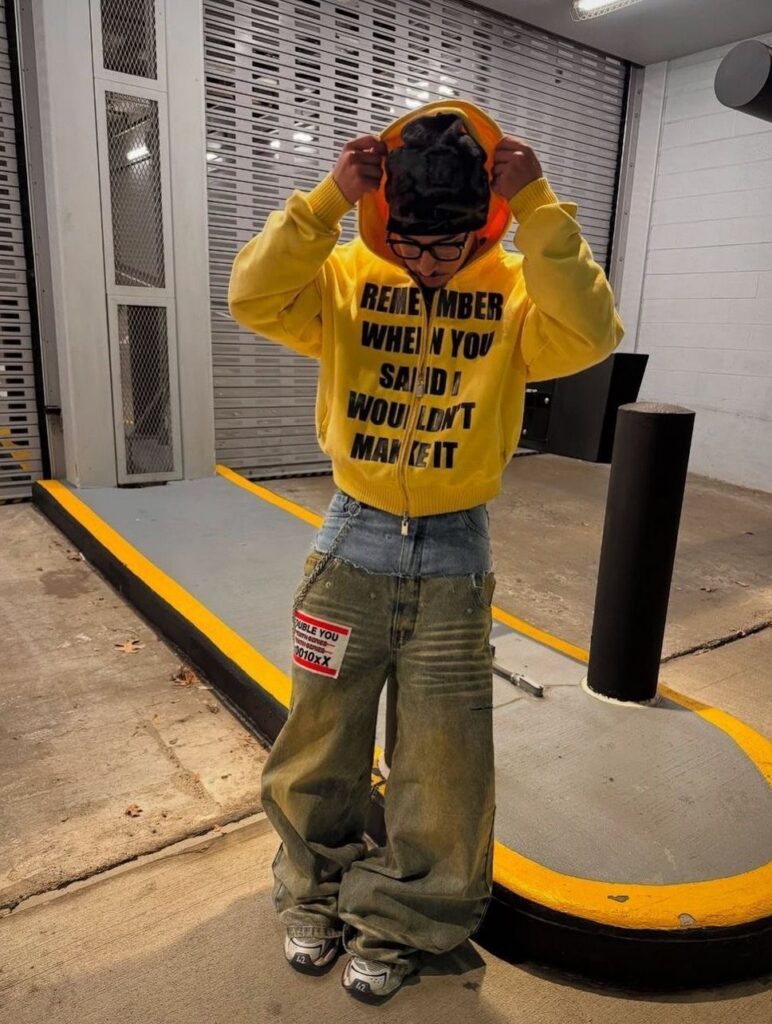 Person in a yellow graphic hoodie and baggy olive trousers standing in an urban concrete area.