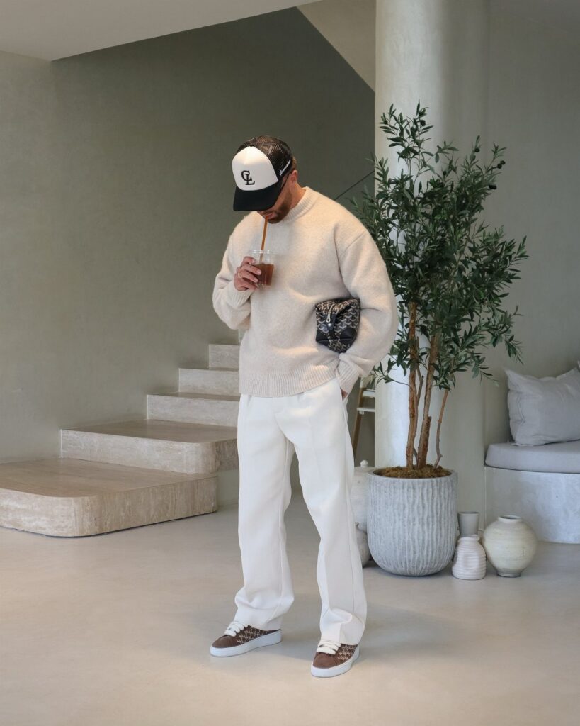 Man in cream sweater and white trousers standing in a minimalist neutral-toned room.