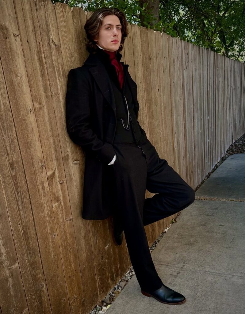 A man in Victorian-style black formal attire leaning against a tall wooden fence outdoors.