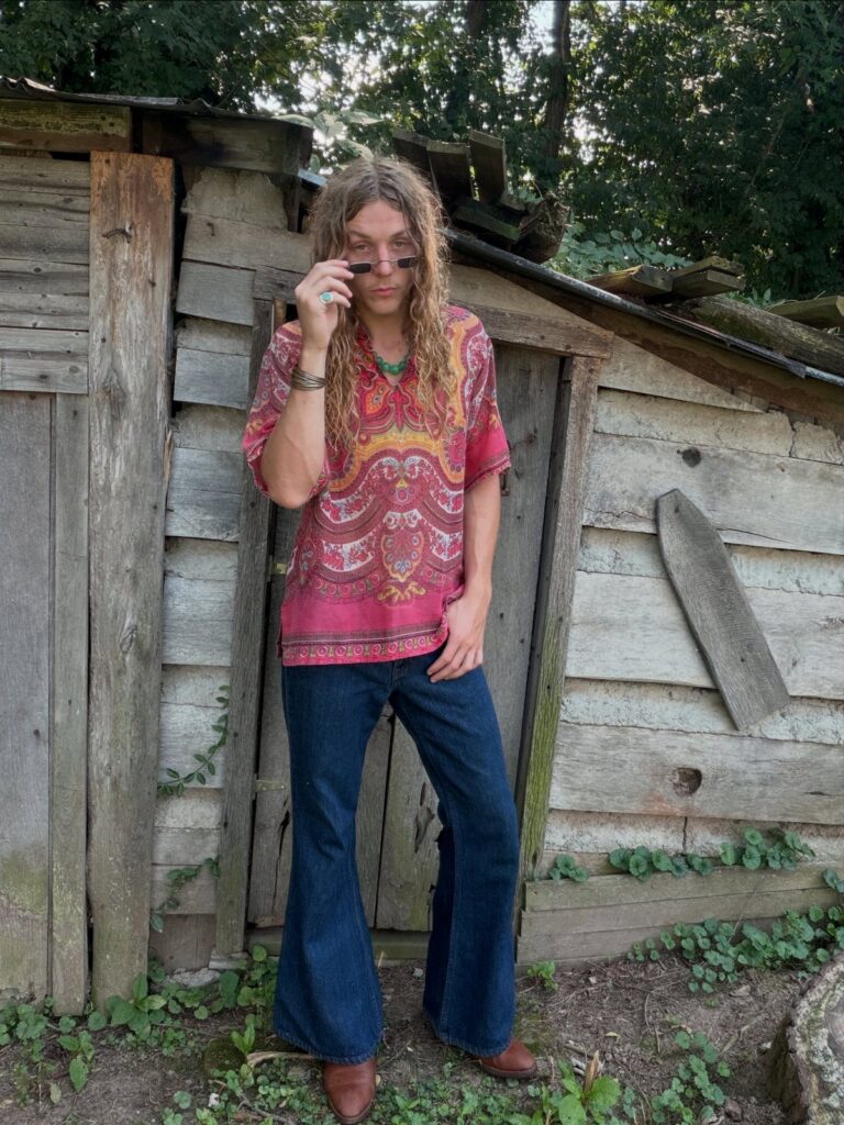 A person wearing a pink patterned tunic and dark blue bell bottom jeans standing outdoors.