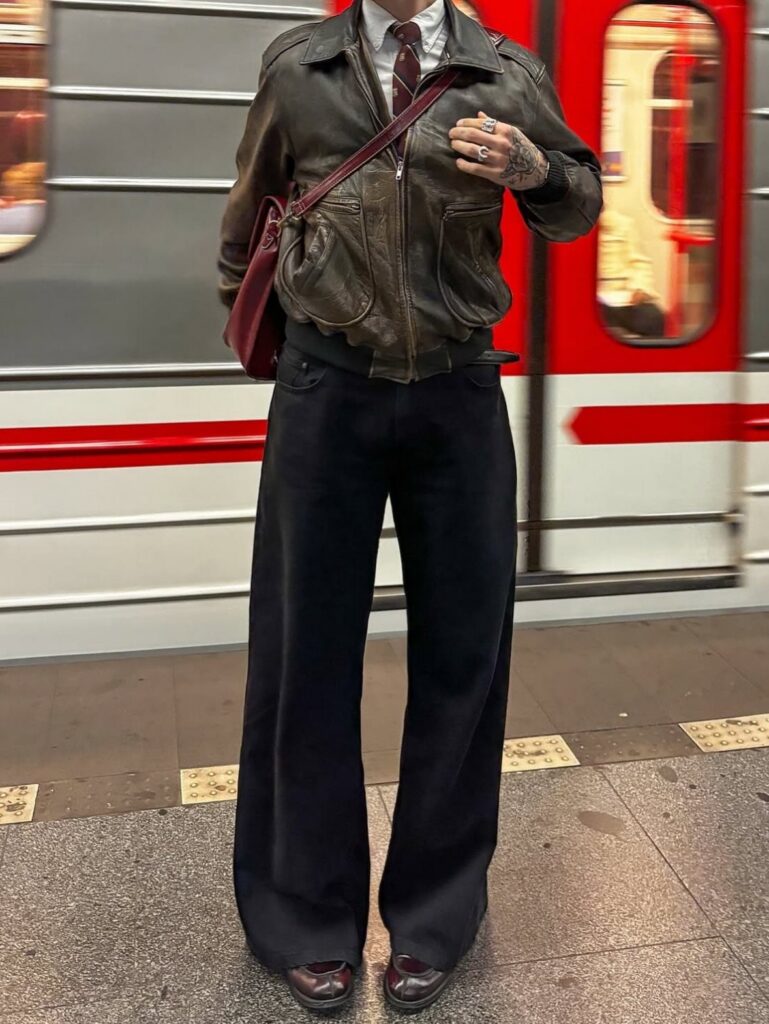 A person dressed in a vintage leather jacket, tie, and wide-leg trousers standing by a subway train.
