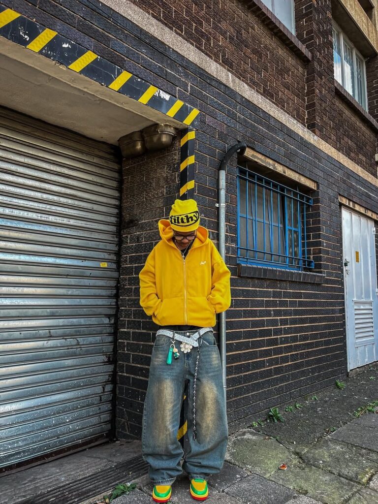 A person wearing a bright yellow hoodie, yellow beanie, and baggy denim jeans standing outdoors.