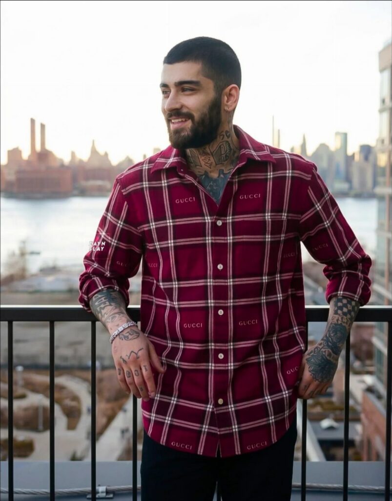 Man with tattoos wearing a red plaid Gucci shirt standing on a balcony overlooking a city.